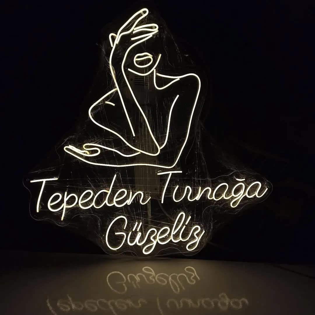 Kadın yüzü çizimi ve 'Tepeden Tırnağa Güzeliz' yazılı sıcak beyaz neon